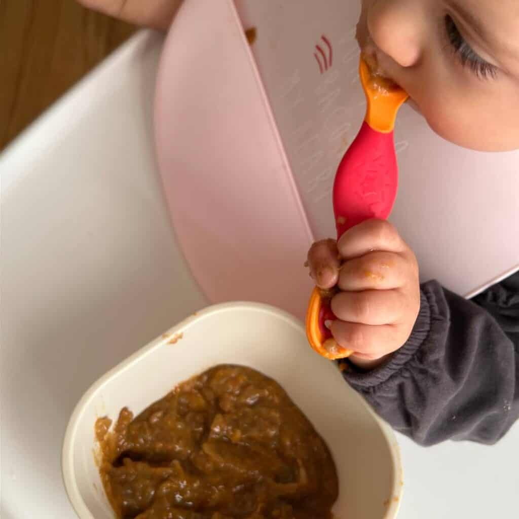 A baby using a spoon to eat blended caldo de lentajas, a traditional Mexican lentil soup.