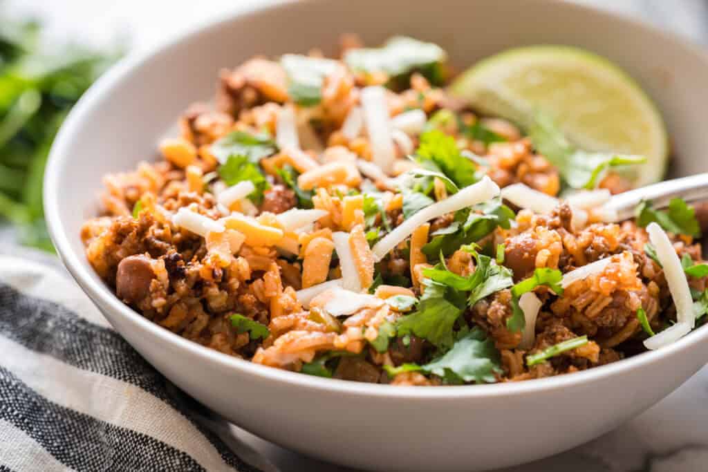 A close-up image of a bowl of sofrito beef beans and rice garnished with cheese, cilantro, and a lime wedge.