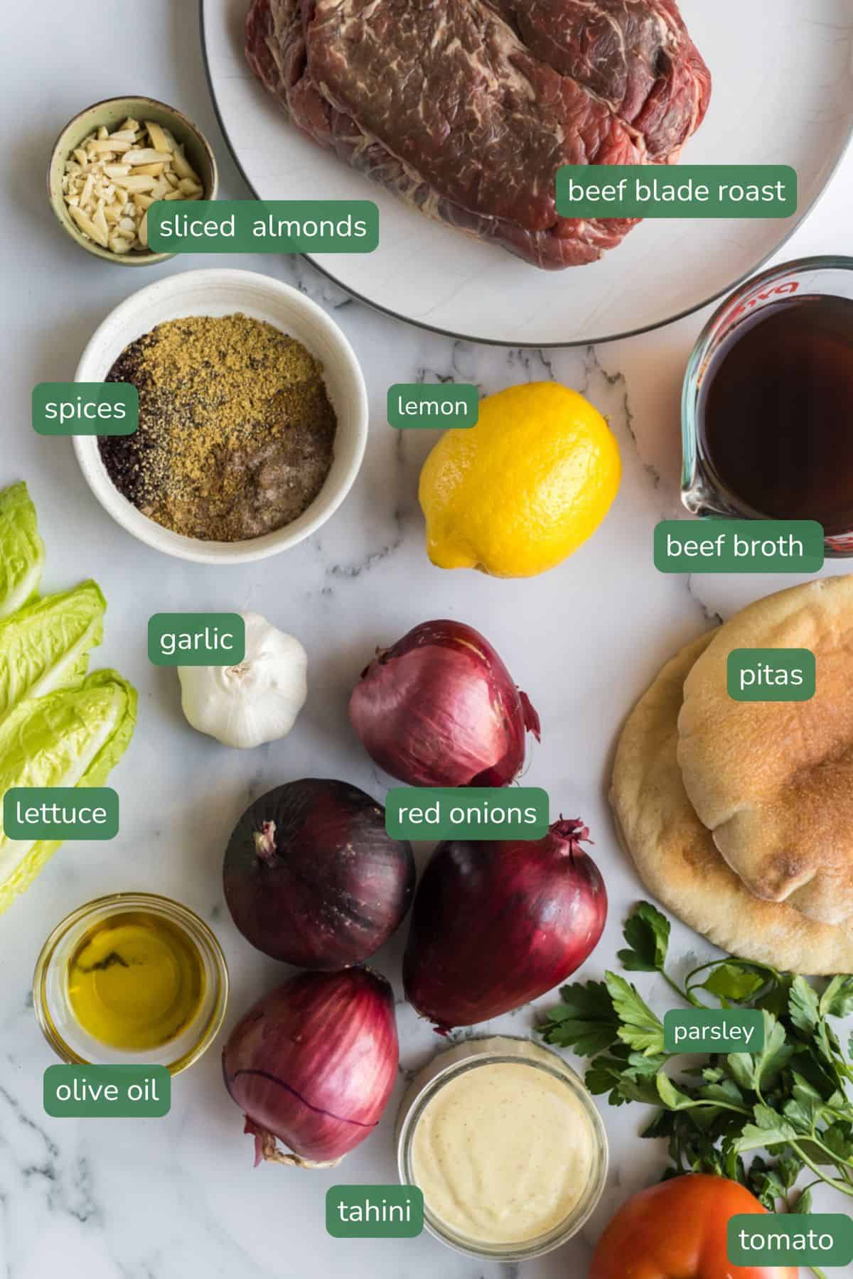 flat lay of ingredients for pull apart sumac beef which includes a beef roast, almonds, spices, lemon, broth, pitas, garlic, red onion, tahini 