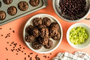 A plate of double chocolate mini muffins, alongside two bowls of the ingredients that make this recipe health boosted - shredded zucchini and black beans!