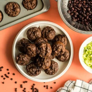 A plate of double chocolate mini muffins, alongside two bowls of the ingredients that make this recipe health boosted - shredded zucchini and black beans!