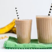 Two glasses of a peanut butter chocolate banana smoothie with bananas and peanuts in the background.