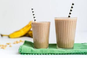 Two glasses of a peanut butter chocolate banana smoothie with bananas and peanuts in the background.