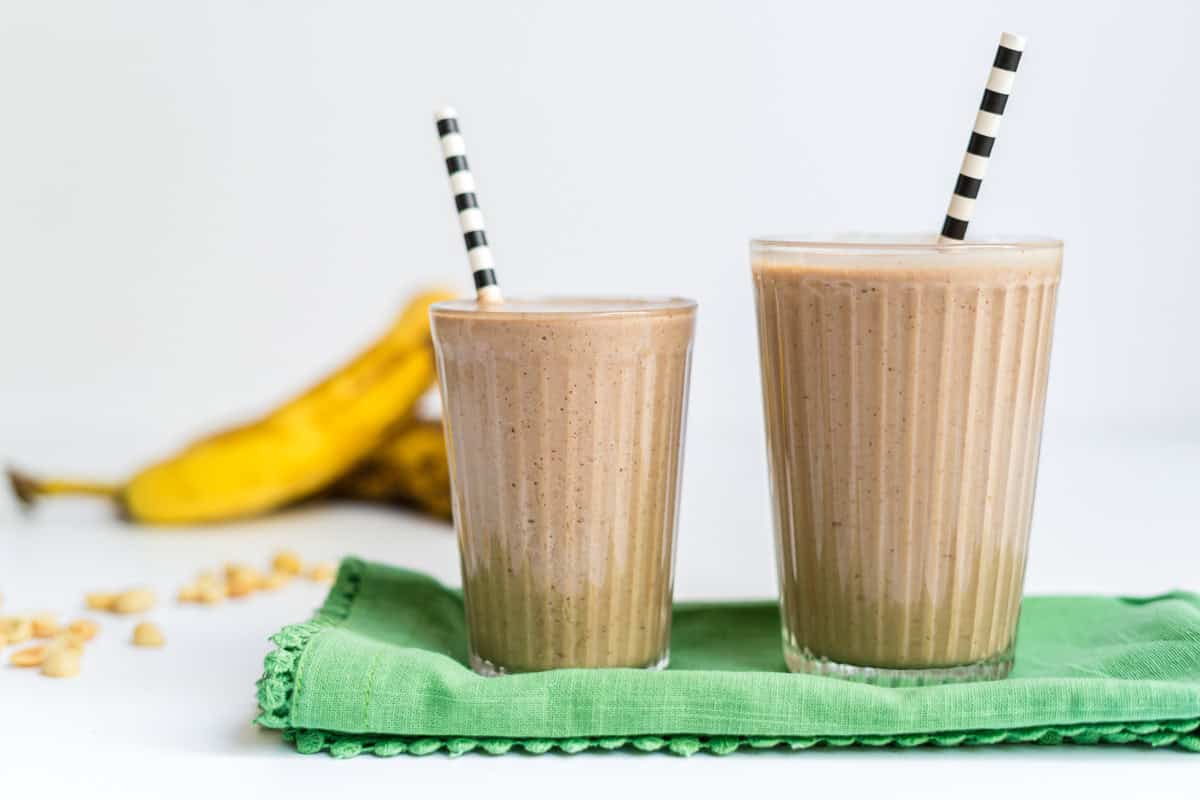 Two glasses of a peanut butter chocolate banana smoothie with bananas and peanuts in the background.