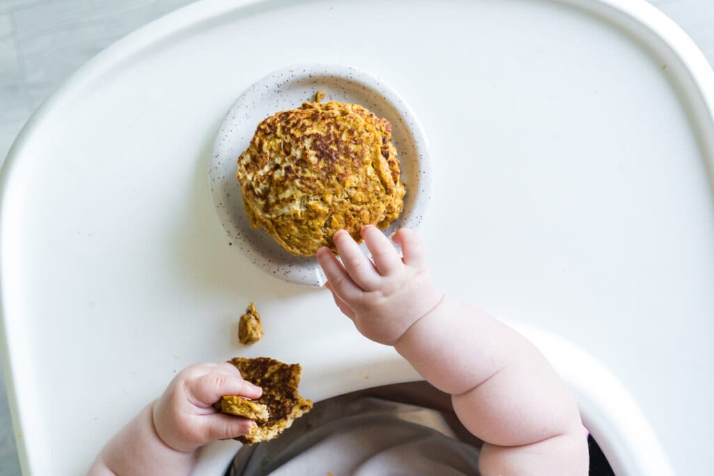 An overhead view of a baby sitting in a high chair reaching for a stack of fruit pouch pancakes.