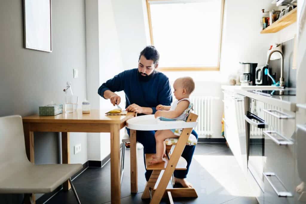 A man is sitting at a table in an apartment kitchen with a baby alongside him in a high chair.