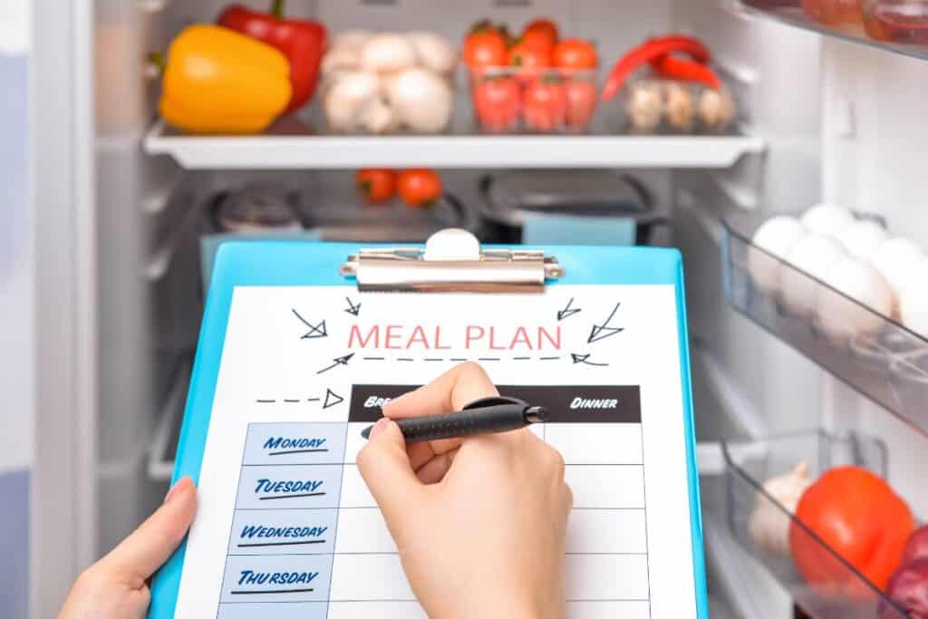 A person is holding a clipboard with a meal planning template in front of an open fridge as part of her meal planning process.