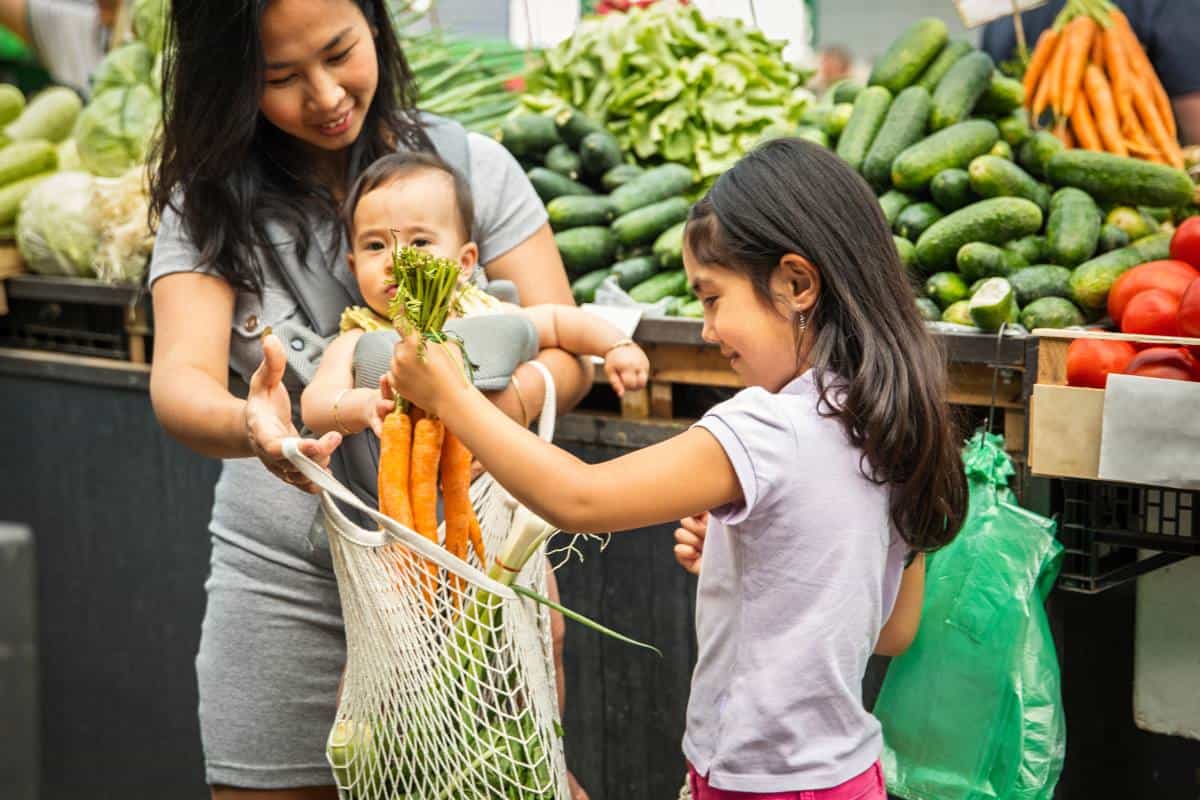 A 10 year old girls puts carrots in a bag while her mom is holding a baby at the grocery store.