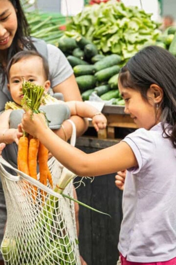A mom holding a baby while her 10 year old daughter puts carrots in a bag at the grocery store.