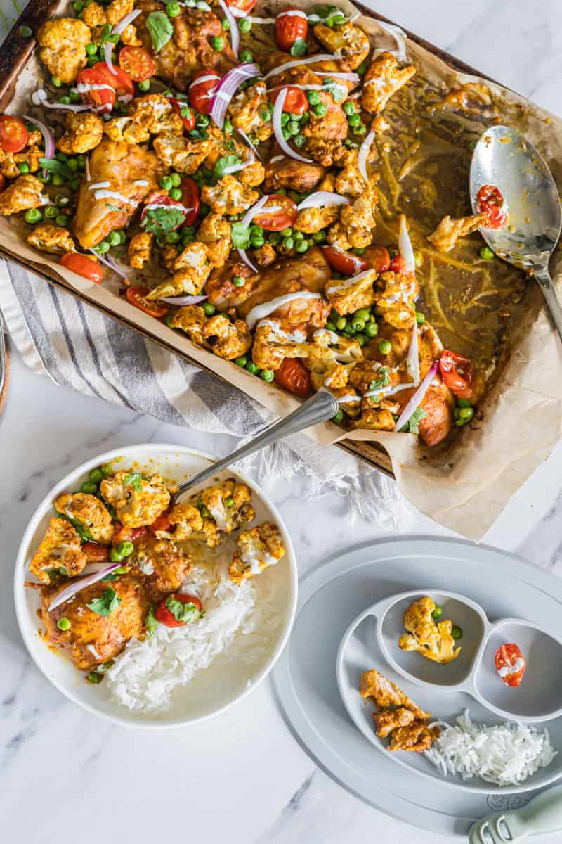 A sheetpan of Indian spiced chicken and veggies beside a plated bowl and a divided plate for a baby.