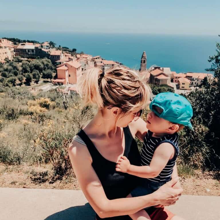 Mama holding toddler in an Italian town with a view of the Mediterranean in the background.