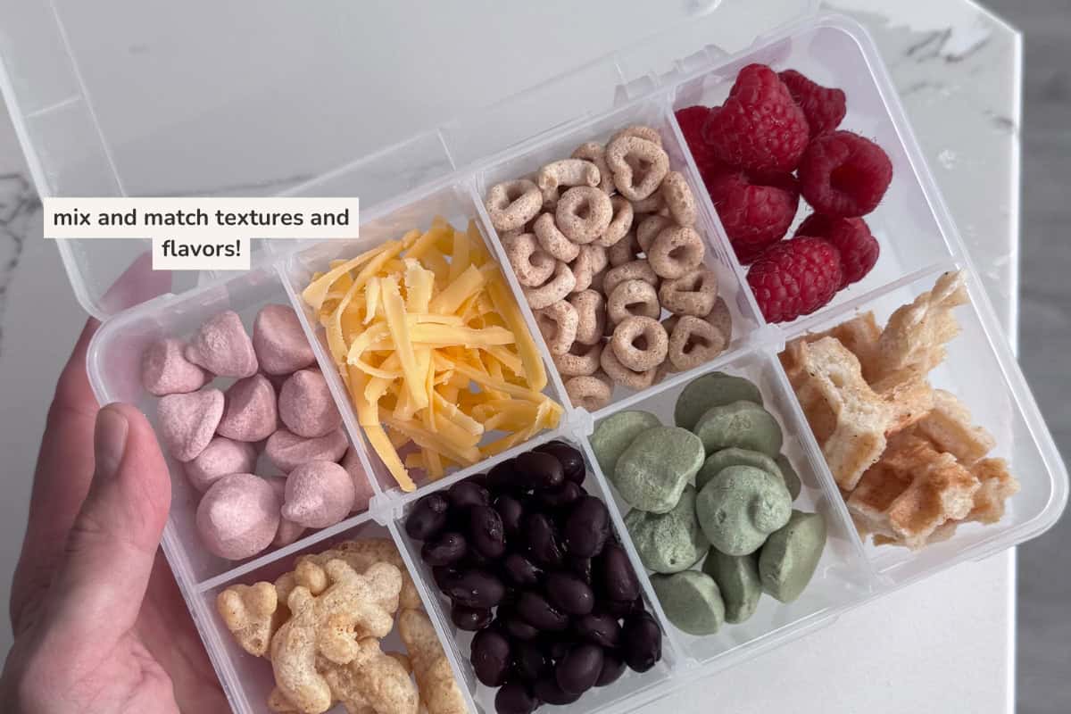 snack box containing small portions yogurt melts, shredded cheese, cereal, berries, waffles, black beans and baby puff snacks