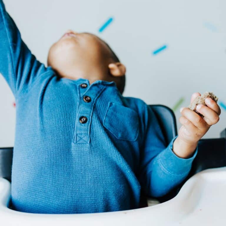 Baby in a high chair arching his back and appears to be throwing or protesting eating food.
