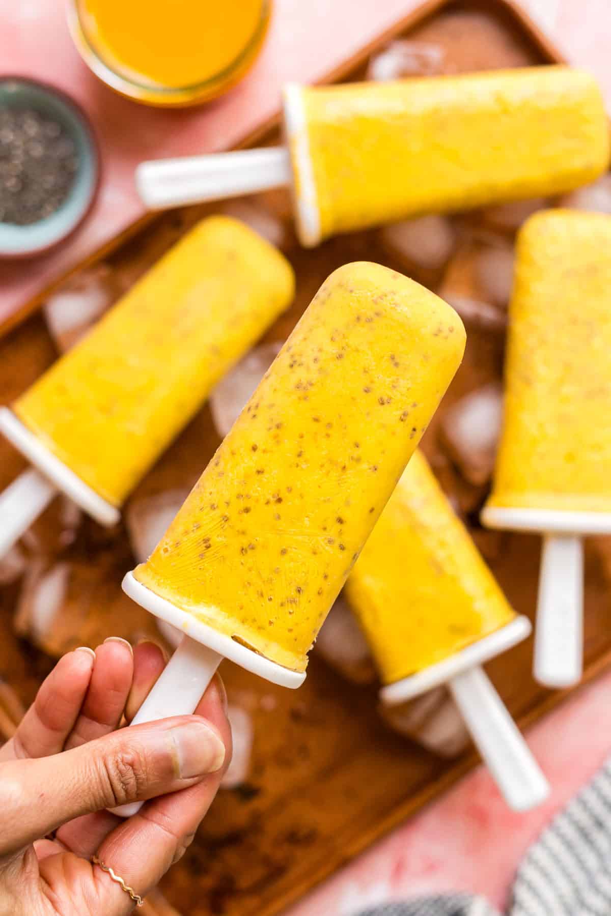 Mango chia popsicles held up by a hand over a tray of other popsicles.