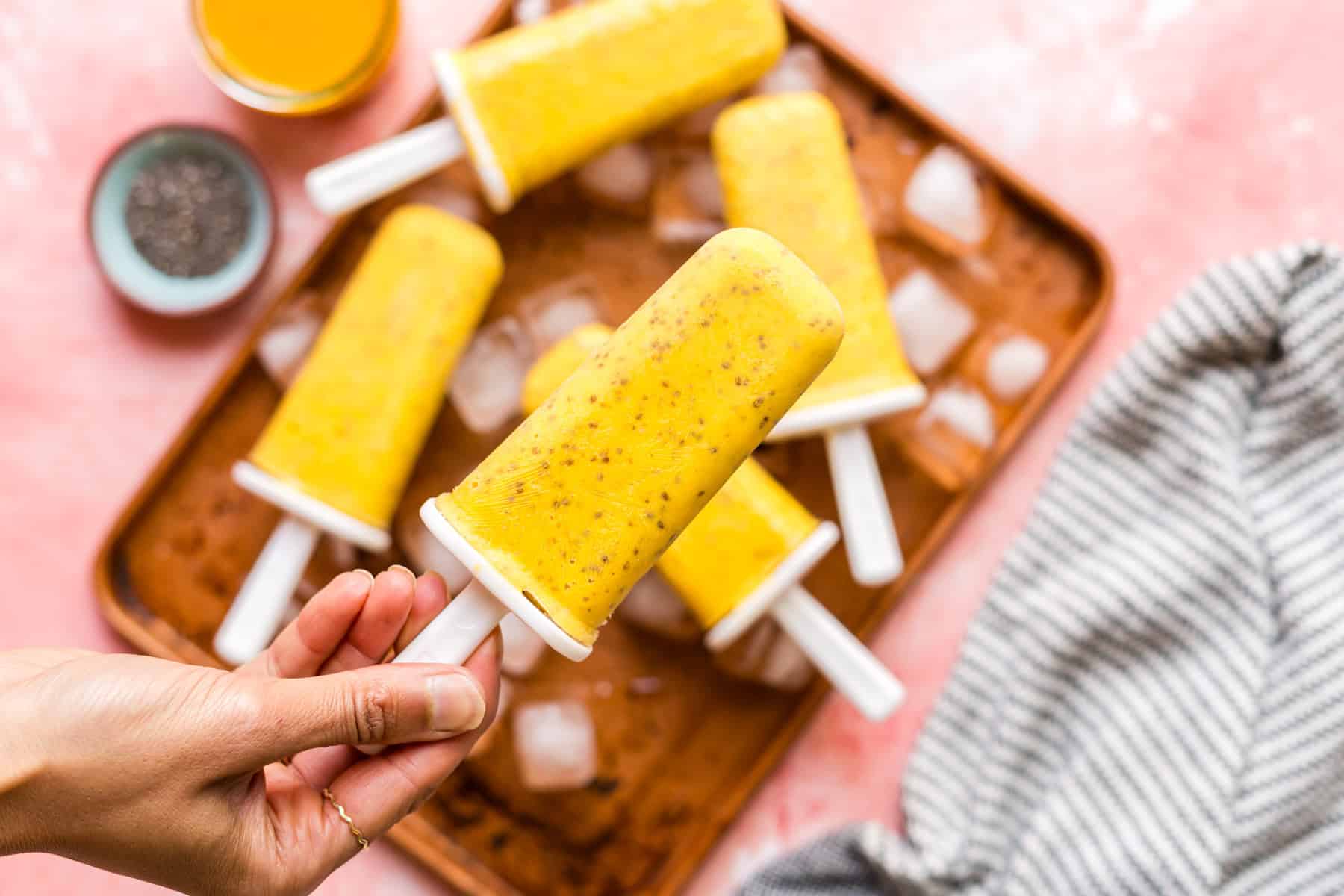 A hand holding a mango chia popsicle with more popsicles in the background.