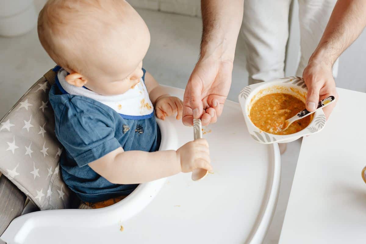baby seated in a highchair holding a spoon. next to baby is a caregiver who also has a bowl with baby food and another spoon.