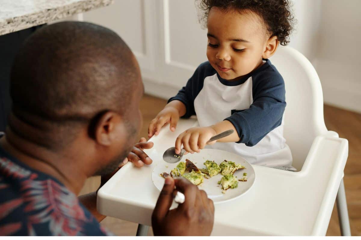 baby seated in a highchair with some sliced avocado toast, across from baby dad is eating too.