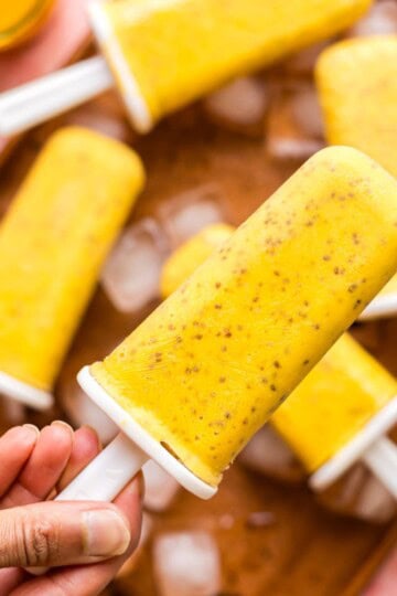 A hand holding a mango chia popsicle in front of a whole batch of popsicles.