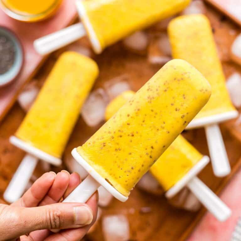 A hand holding a mango chia popsicle in front of a whole batch of popsicles.