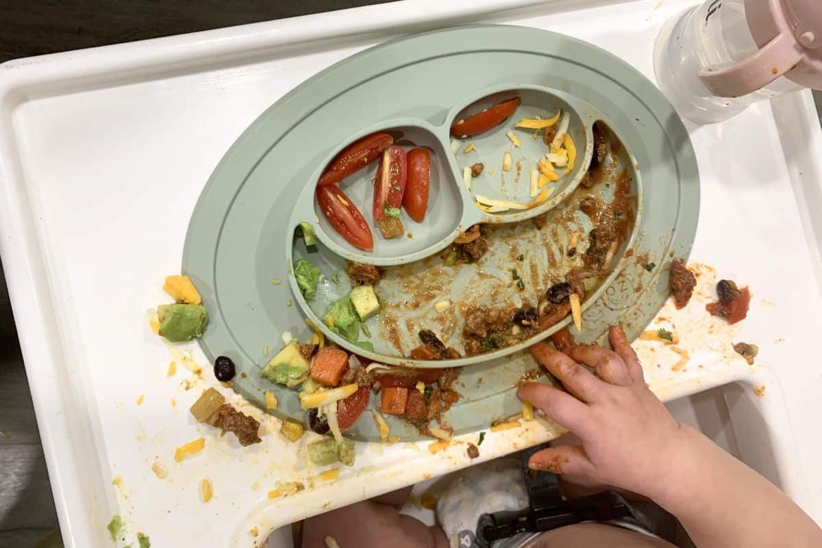 EZPZ branded plate with food that appears to be very messy on a highchair. There are also small baby hands over the food.