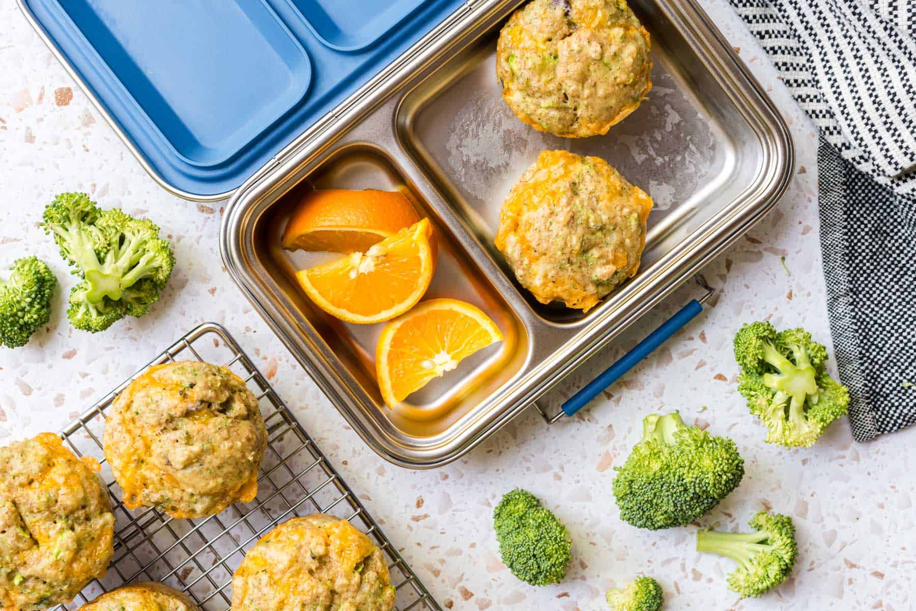 A stainless steel lunchbox with two fluffy cheddar egg muffins and orange slices beside a cooling rack of more fluffy cheddar egg muffins.