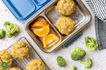 A stainless steel lunchbox with two fluffy cheddar egg muffins and orange slices beside a cooling rack of more fluffy cheddar egg muffins.