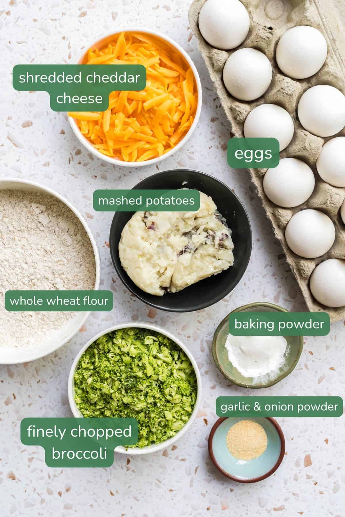 Flat lay of ingredients for cheddar egg muffins--a carton of eggs, shredded cheddar cheese, mashed potatoes, whole wheat flour, shredded broccoli, baking powder and garlic powder.