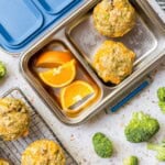 A stainless steel lunchbox with two fluffy cheddar egg muffins and orange slices beside a cooling rack of more fluffy cheddar egg muffins.