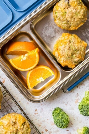 A stainless steel lunchbox with two fluffy cheddar egg muffins and orange slices beside a cooling rack of more fluffy cheddar egg muffins.