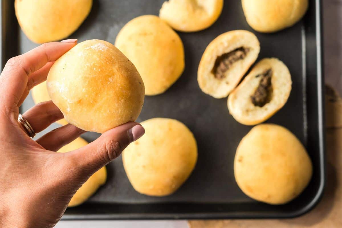 Hand holding up a baked mennonite meat bun with other meatbuns in the background on a baking sheet.