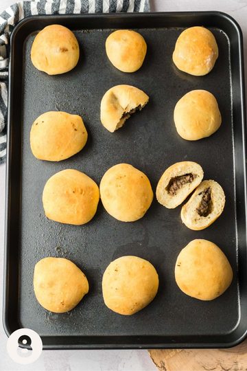 Baked and cooked mennonite meat buns arranged on a baking sheet--one of the meatbuns has been sliced through to showcase the filling and cross-section.