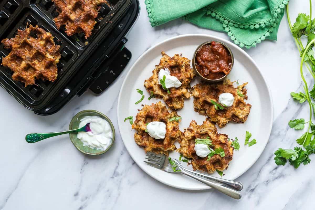 A plate with four chicken taco waffles laid beautifully and topped with sour cream and cilantro as a garnish. Next to the waffle is a little bowl with salsa as a dipping sauce. In the image is also a waffle iron with two taco waffles inside. 