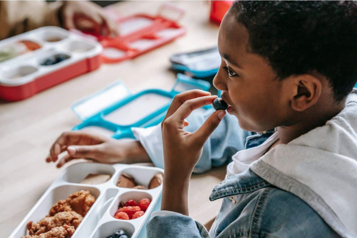 A boy sitting at a lunch table with a packed lunchbox in front of him. He is bringing a blueberry to his mouth.